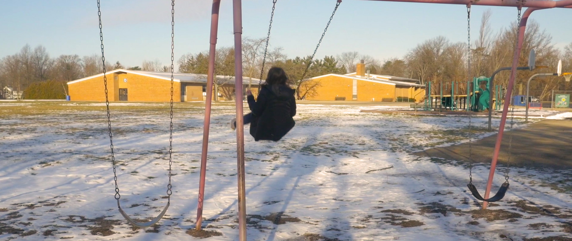 Jordyn on a swing seen from the back, in a school yard field covered with snow 